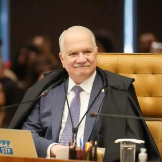 Homem idoso, juiz ou autoridade jurídica, sentado em tribunal, usando toga e vestimenta formal, com pessoas ao fundo, simbolizando ambiente de julgamento ou sessão de tribunal, Direito Hoje Notícias, foco em justiça e direito.