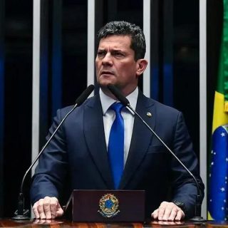 Legislador brasileiro em discurso no plenário durante sessão parlamentar, com bandeira do Brasil ao fundo, voltado para temas de direito e política no Brasil.