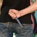 Facas e ameaças: homem com uma faca na mão atrás das costas, em um ambiente com parede grafitada, representando temas de segurança, violência e direito na atualidade.