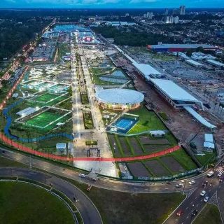 Praça de eventos e lazer com áreas verdes, pistas de caminhada, esportes e espaço para convivência na cidade ao entardecer, iluminada com teto de luzes e construções modernas em fundo urbano.