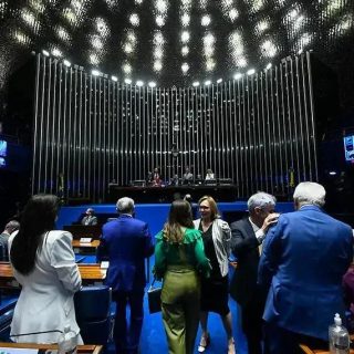 Alo histórico de uma sessão de tribunal ou assembleia legislativa com pessoas conversando, incluindo mulheres e homens, ambiente formal e moderno com iluminação e telas digitais ao fundo.