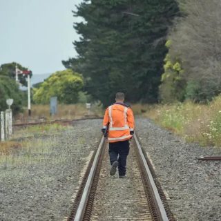 Homem de costas caminhando sobre trilhos de trem em área rural, vestindo jaqueta laranja, representando segurança e prevenção em operações ferroviárias.