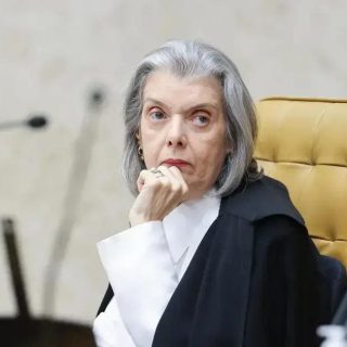 Juíza de tribunal em audiência judicial, vestindo toga preta, com expressão séria e pensativa, diante de bancada com microfones e documentos, em ambiente de tribunal ou fórum.