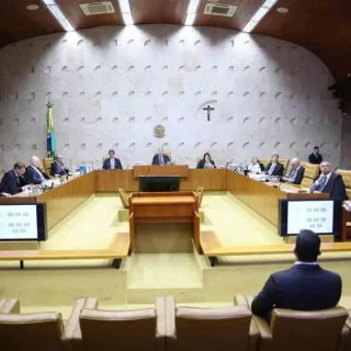 Pessoas sentadas em reunião do tribunal, discutindo temas jurídicos e sobre direito, em ambiente formal e oficial, na sala de audiência do judiciário brasileiro, com destaque para a bandeira e símbolos nacionais.