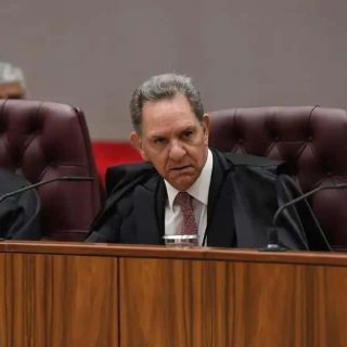 Juiz do tribunal durante sessão judicial com profissionais do direito e magistrados ao fundo.