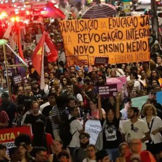Discurso de protesto exige melhorias na educação pública, com cartazes e faixas pedindo revogação de medidas que prejudicam o ensino médio, destacando mobilização de estudantes e apoiadores por direitos educacionais.