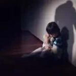 Menina triste e solitária sentada no chão com sombra de uma figura ameaçadora ao fundo, simbolizando vulnerabilidade e risco para crianças.