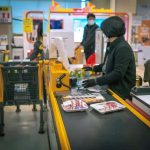 Carnê de supermercado com caixas registradoras e clientes no checkout, focando em compras de alimentos, produtos de higiene e promoções em uma loja moderna.